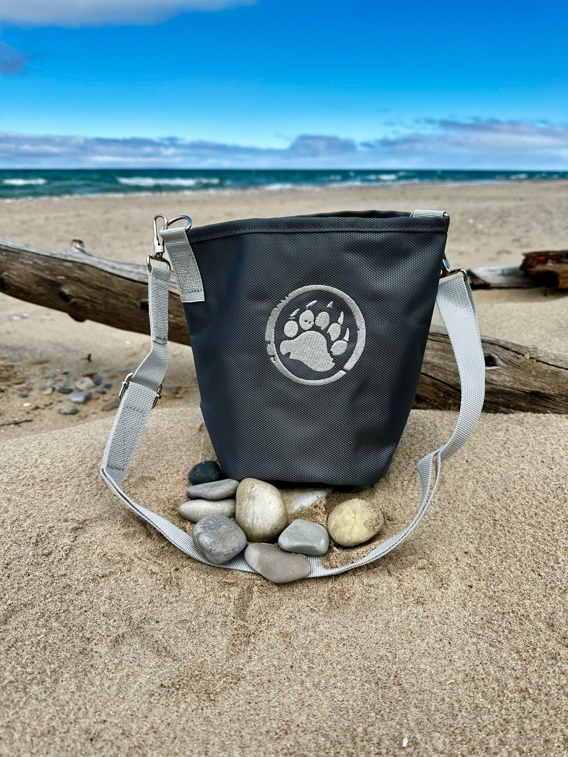 Black bag with a paw print logo on a sandy beach with stones and driftwood.