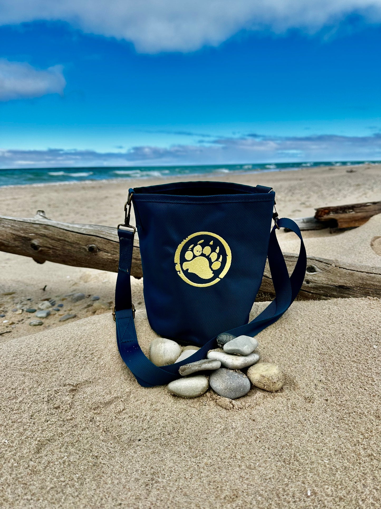 Navy blue bag with a paw print logo on a sandy beach with pebbles and driftwood.