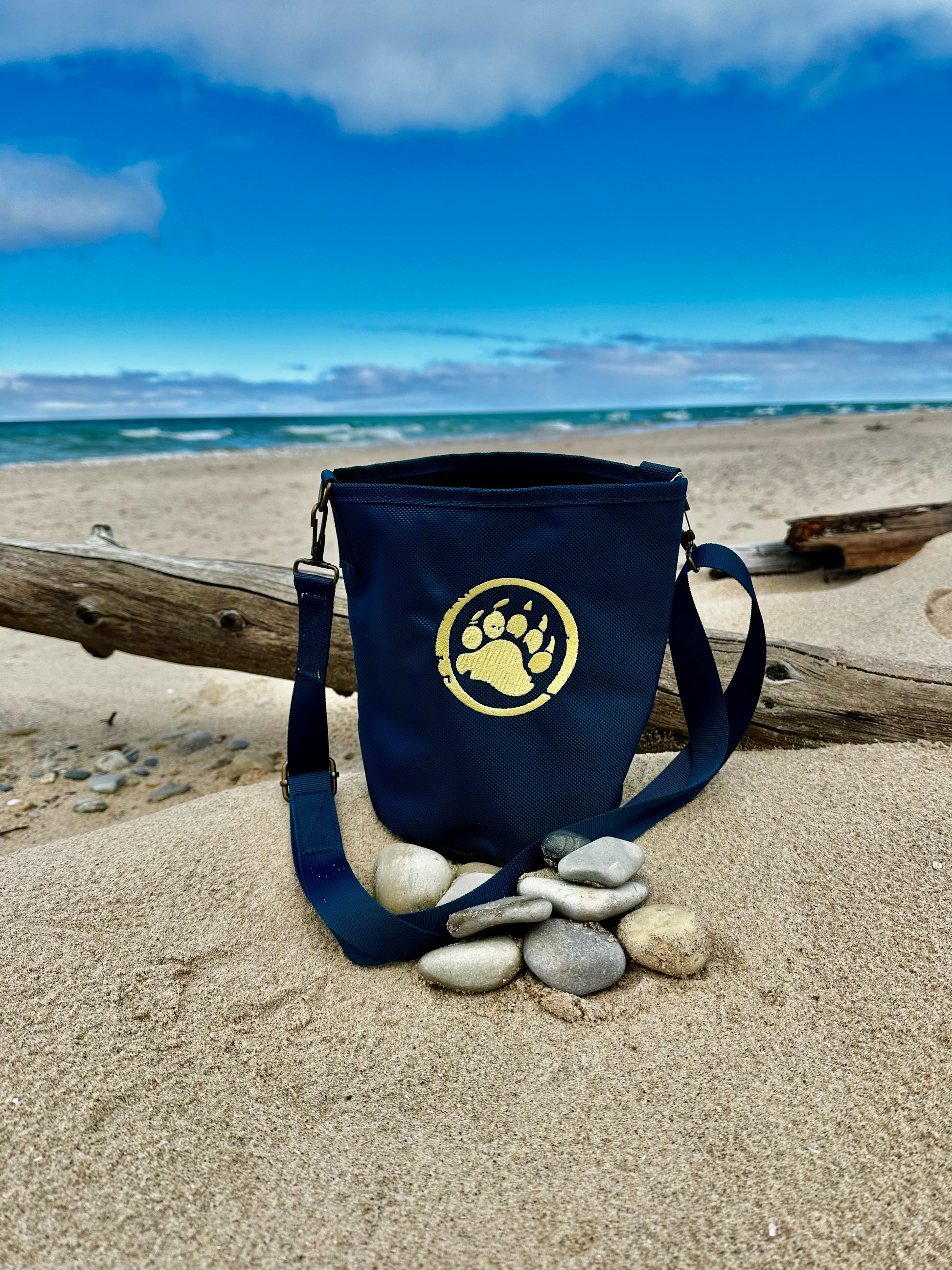 Navy blue bag with a paw print logo on a sandy beach with pebbles and driftwood.